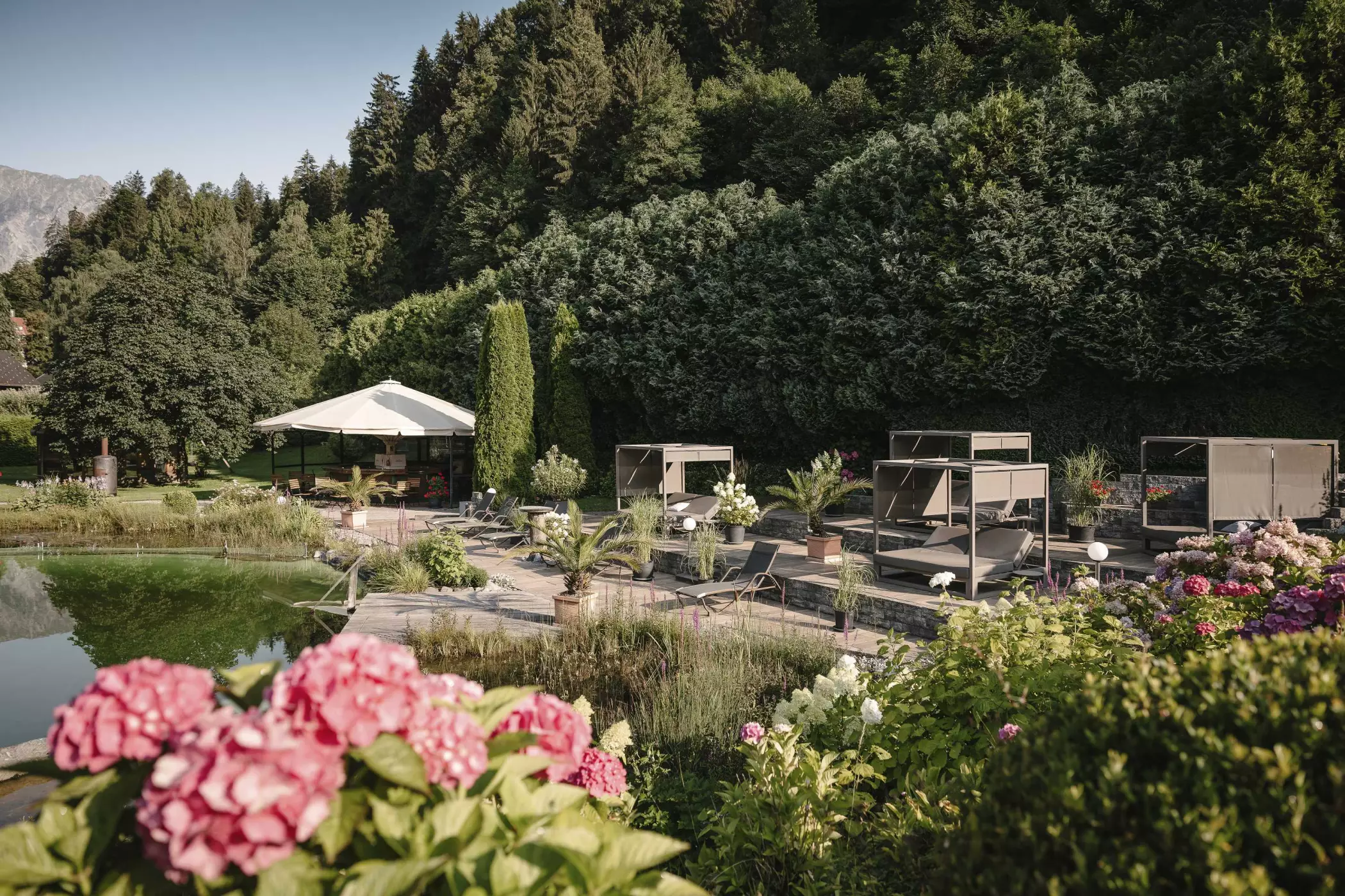 Gartenbereich mit Naturpool, blühenden Hortensien, Sonnenliegen und überdachten Daybeds vor einem Hintergrund aus Bäumen und Bergen.